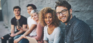 A group of smiling university students is sitting outside of a building.