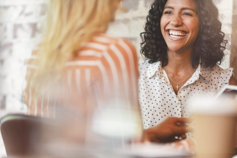 Two happy women are smiling and talking in a coffee shop.