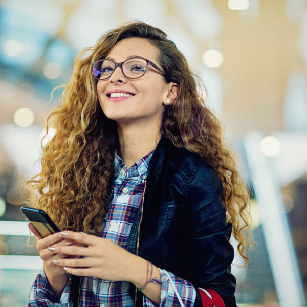 Young adult woman is smiling and holding her smartphone, as she just had a great customer experience.